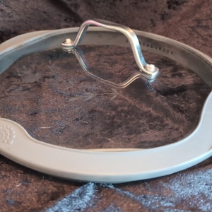 May include: A clear glass lid with a silver handle and a light gray rim. The lid is round and appears to be for a pot or pan. The handle is arched and attached to the glass with small silver rivets. The rim has a decorative embossed design.