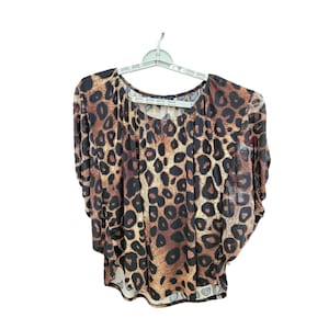 May include: A leopard print top with short sleeves hangs on a white hanger. The top features a brown and black spotted pattern. The fabric appears lightweight and flowing, with a gathered neckline and a slightly asymmetrical hemline.