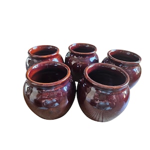 May include: A set of five small, round ceramic pots with a glossy, deep burgundy glaze. Each pot features a wide, open mouth with a slightly flared rim and a smooth, rounded body. The dark red finish has subtle variations in tone and light reflection.