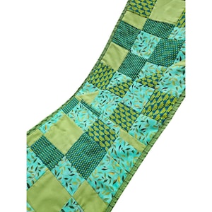 May include: A rectangular table runner featuring a patchwork design of alternating fabric squares in shades of lime green, teal, and dark green. The pattern includes solid colors, small yellow polka dots, fan-like motifs, and green leaf prints.