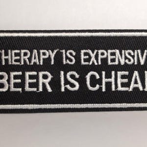 Op de afbeelding: Zwart rechthoekig embleem met wit geborduurde tekst: "THERAPY IS EXPENSIVE BEER IS CHEAP". Het embleem heeft een witte rand.