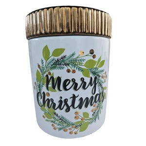 May include: A white ceramic container with a gold-colored lid. The container is decorated with a green wreath and the words "Merry Christmas" in black script. The wreath includes green leaves and small berries.