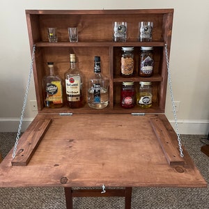 May include: A wooden bar cabinet with a fold-down top and a chain to secure it. The cabinet has shelves with various bottles and jars, including liquor, jars of jam, and shot glasses.
