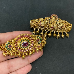 Peut inclure: Deux barrettes dorées avec des motifs complexes et des pierres précieuses rouges et vertes. La barrette du haut présente une figure de divinité et des éléphants, tandis que la barrette du bas a un motif floral.