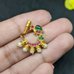 Op de afbeelding: Een goudkleurige neusring met een gebogen ontwerp. Het heeft een groene edelsteen, roze en groene accenten en kleine witte parels. De neusring heeft een schroefachtige paal voor veilig dragen.