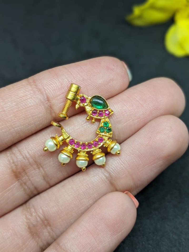 Op de afbeelding: Een goudkleurige neusring met een gebogen ontwerp. Het heeft een groene edelsteen, roze en groene accenten en kleine witte parels. De neusring heeft een schroefachtige paal voor veilig dragen.