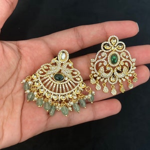 May include: Gold-toned earrings with intricate detailing. Each earring features a central green gemstone, surrounded by clear crystals. One earring has a peacock design, while the other has a floral pattern. Both have dangling green beads and small white pearls.