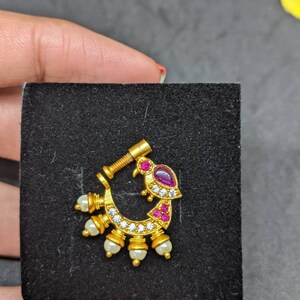 Op de afbeelding: Een goudkleurige neusring met een gebogen ontwerp. Het heeft een vogelachtige vorm met een paarse edelsteen en rode accenten. De ring is versierd met kleine witte parels en heldere edelstenen. De neusring wordt gepresenteerd op een zwart oppervlak.
