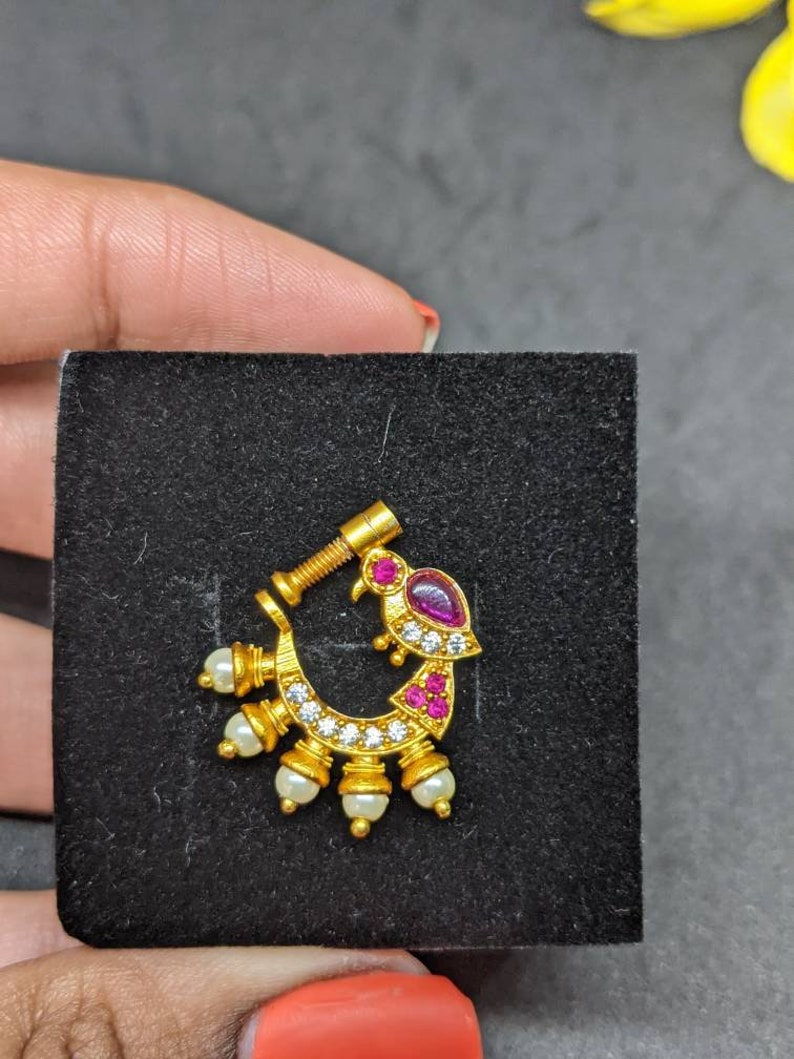Op de afbeelding: Een goudkleurige neusring met een gebogen ontwerp. Het heeft een vogelachtige vorm met een paarse edelsteen en rode accenten. De ring is versierd met kleine witte parels en heldere edelstenen. De neusring wordt gepresenteerd op een zwart oppervlak.