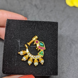 Op de afbeelding: Een goudkleurige neusring met een pauwontwerp. De sieraden hebben een gebogen vorm met een rij kleine witte parels en heldere stenen. De pauw heeft groene, rode en heldere stenen accenten. De neusring wordt op een zwart oppervlak gepresenteerd.