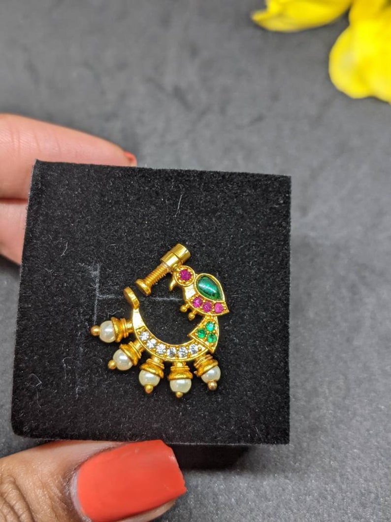 Op de afbeelding: Een goudkleurige neusring met een pauwontwerp. De sieraden hebben een gebogen vorm met een rij kleine witte parels en heldere stenen. De pauw heeft groene, rode en heldere stenen accenten. De neusring wordt op een zwart oppervlak gepresenteerd.