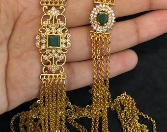 Op de afbeelding: Een goudkleurige ketting met meerdere strengen en twee verschillende decoratieve middenstukken. Het ene middenstuk is een langwerpige filigraanplaat met een vierkante groene steen, het andere is een cirkelvormig cluster met een ovale groene steen.