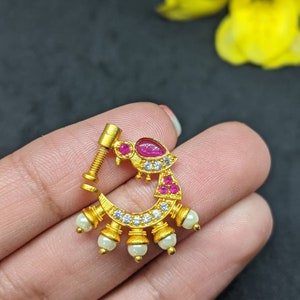 Op de afbeelding: Een goudkleurige neusring met een schroefpost. De ring heeft een rode edelsteen, kleine witte edelstenen en bungelende witte parels. Het ontwerp is sierlijk en gedetailleerd, met de nadruk op de edelstenen en parels.