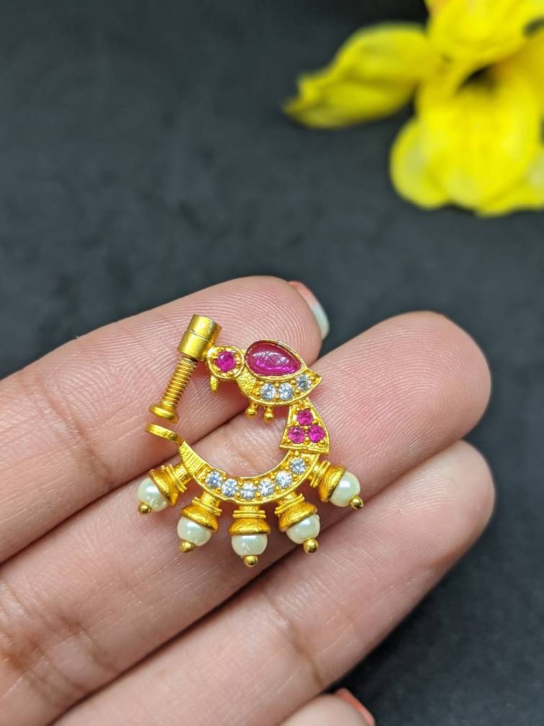 Op de afbeelding: Een goudkleurige neusring met een schroefpost. De ring heeft een rode edelsteen, kleine witte edelstenen en bungelende witte parels. Het ontwerp is sierlijk en gedetailleerd, met de nadruk op de edelstenen en parels.