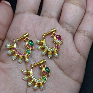 Op de afbeelding: Drie goudkleurige neusringen met een halve maanvorm. Elke ring heeft een ontwerp met rode, groene en witte edelstenen, samen met kleine witte parels. De ringen worden gepresenteerd op een lichtbruin oppervlak.