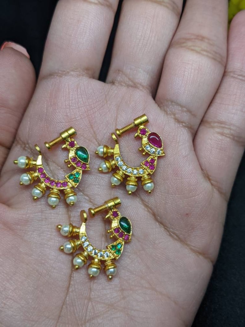 Op de afbeelding: Drie goudkleurige neusringen met een halve maanvorm. Elke ring heeft een ontwerp met rode, groene en witte edelstenen, samen met kleine witte parels. De ringen worden gepresenteerd op een lichtbruin oppervlak.