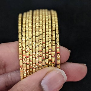 May include: A stack of 16 gold-tone bangles with a textured design. The bangles are stacked on top of each other and are held in a hand.