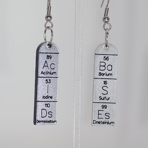 May include: A pair of silver earrings with a chemical element design. The left earring features the element Actinium (Ac), Iodine (I), and Darmstadtium (Ds). The right earring features the element Barium (Ba), Sulfur (S), and Einsteinium (Es).