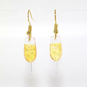Puede incluir: Un par de pendientes de aro dorados con un dije de copa de champán de vidrio transparente. La copa está llena de un líquido amarillo que parece champán.