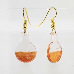 Könnte beinhalten: Ein Paar goldene Ohrhänger mit klaren Glasfläschchen, gefüllt mit kupferfarbenem Glitzer.