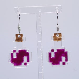 May include: A pair of pixelated potion bottle earrings. The earrings feature a clear, square-shaped bottle design with a cork top and a vibrant magenta liquid inside. The earrings hang from silver-colored hooks.