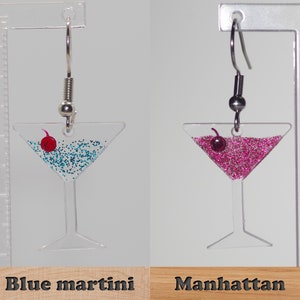 May include: A pair of dangle earrings featuring a martini glass filled with blue glitter and a red cherry, and a martini glass filled with pink glitter and a red cherry. The earrings are labeled "Blue martini" and "Manhattan".