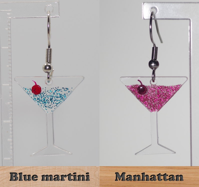 May include: A pair of dangle earrings featuring a martini glass filled with blue glitter and a red cherry, and a martini glass filled with pink glitter and a red cherry. The earrings are labeled "Blue martini" and "Manhattan".