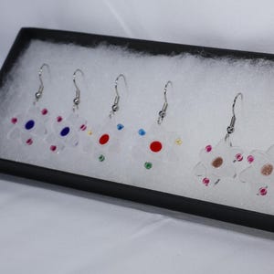 Może przedstawiać: Zestaw przezroczystych kolczyków w kształcie kwiatów z akrylu ze srebrnymi haczykami, prezentowany w czarnym pudełku. Każdy kolczyk ma kolorowe akcenty, w tym czerwony, niebieski i złoty, oraz małe, kolorowe klejnoty. Kolczyki mają motyw kwiatowy.