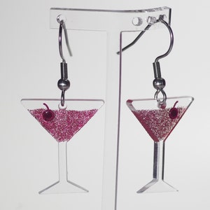 May include: A pair of silver dangle earrings with a clear glass martini glass charm filled with pink glitter and a red cherry on top.
