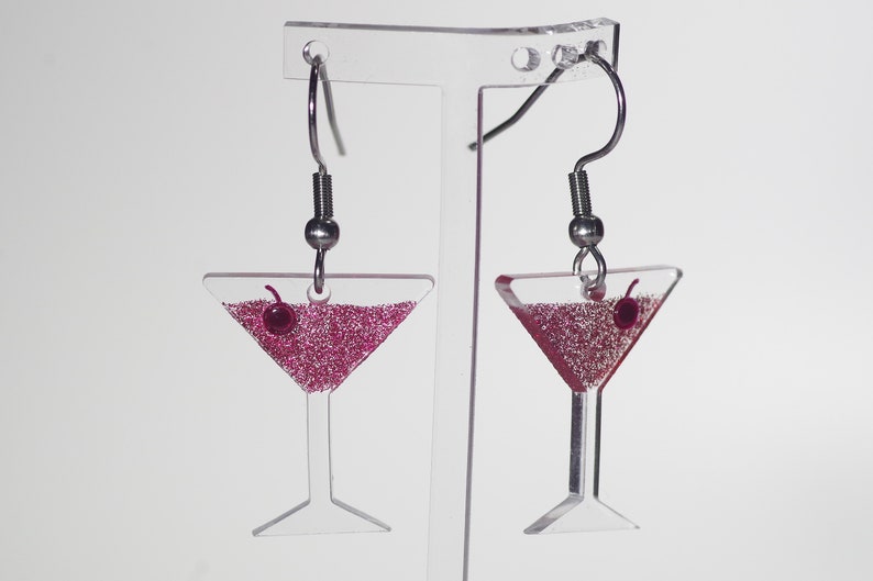 May include: A pair of silver dangle earrings with a clear glass martini glass charm filled with pink glitter and a red cherry on top.