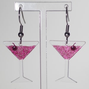 May include: A pair of clear acrylic earrings shaped like martini glasses filled with pink glitter and a red cherry on top. The earrings have silver hooks.