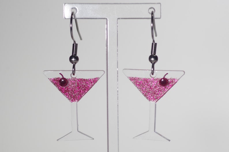 May include: A pair of clear acrylic earrings shaped like martini glasses filled with pink glitter and a red cherry on top. The earrings have silver hooks.