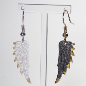 May include: A pair of dangle earrings with gold accents. One earring features a white wing with gold glitter, and the other earring features a black wing with gold glitter.