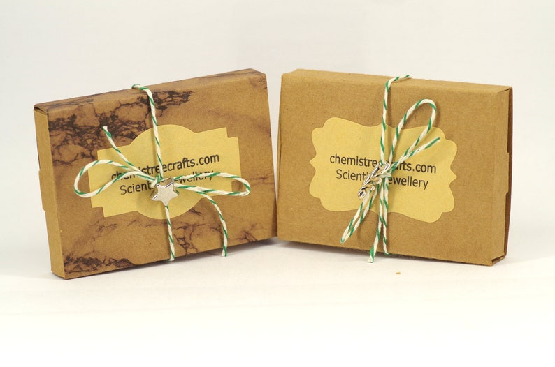 May include: Two small, brown cardboard boxes tied with twine and a green and white ribbon. The boxes have labels with the text "chemistrecrafts.com Scientific Jewellery".