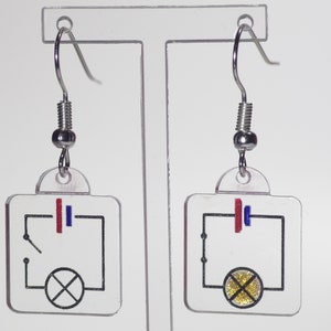 Puede incluir: Un par de pendientes de plata con un dije cuadrado transparente que presenta un diagrama de circuito simple con una bombilla y una batería. La bombilla es de purpurina dorada.
