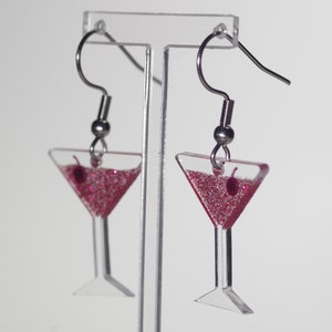May include: A pair of silver dangle earrings with pink glitter martini glass charms. The charms have a small red cherry on the rim.
