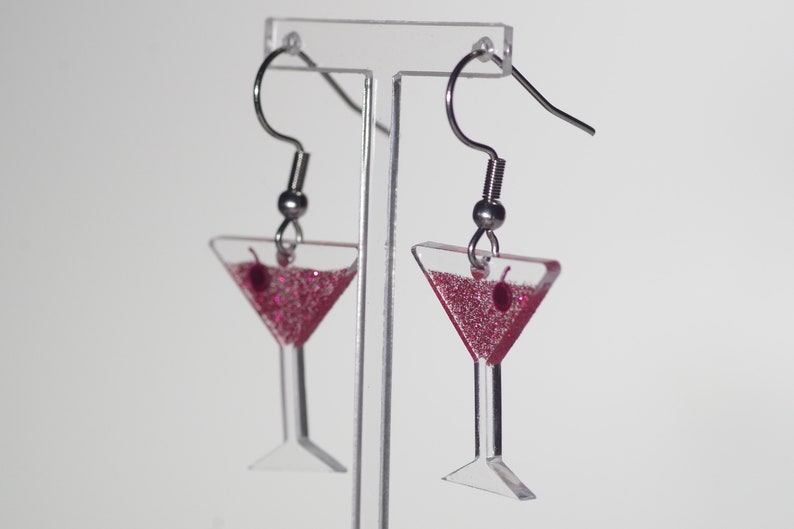 May include: A pair of silver dangle earrings with pink glitter martini glass charms. The charms have a small red cherry on the rim.