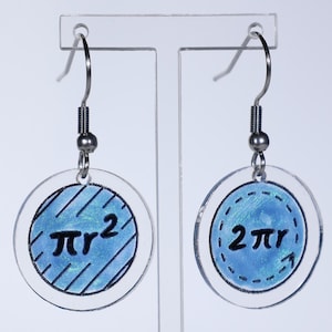 Puede incluir: Un par de pendientes de acrílico transparente con ganchos plateados. Cada pendiente presenta un diseño circular con un centro azul y texto negro. Uno muestra "πr²" y el otro "2πr".