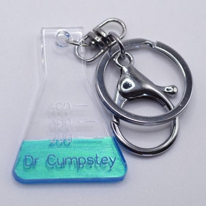 May include: A clear plastic beaker keychain with a blue and green liquid inside. The beaker has markings for 200, 300, and 400. The keychain has a silver metal ring and a clasp. The text "Dr. Cumpstey" is printed on the beaker.