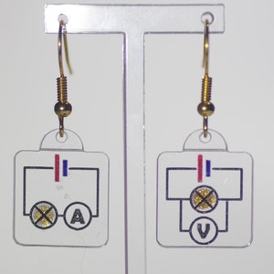 Puede incluir: Un par de pendientes de oro con dijes de plástico transparente que representan diagramas de circuitos eléctricos simples. Un dije tiene una bombilla y la letra 'A' para Amperaje. El otro dije tiene una bombilla y la letra 'V' para Voltaje.