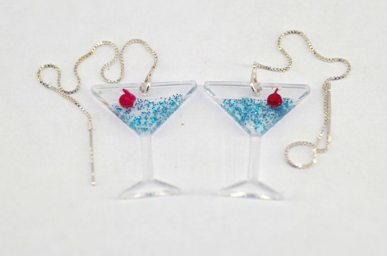 May include: A pair of silver chain earrings with a clear glass martini glass charm filled with blue glitter and a red cherry.