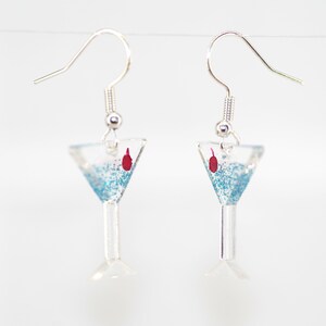 May include: A pair of silver dangle earrings with a blue glitter martini glass charm and a red cherry on top.
