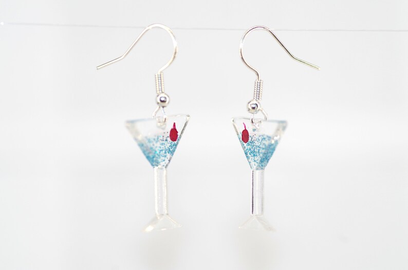 May include: A pair of silver dangle earrings with a blue glitter martini glass charm and a red cherry on top.