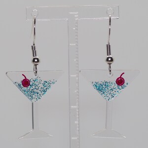 May include: A pair of silver dangle earrings with a clear glass martini glass charm filled with blue glitter and a red cherry on top.