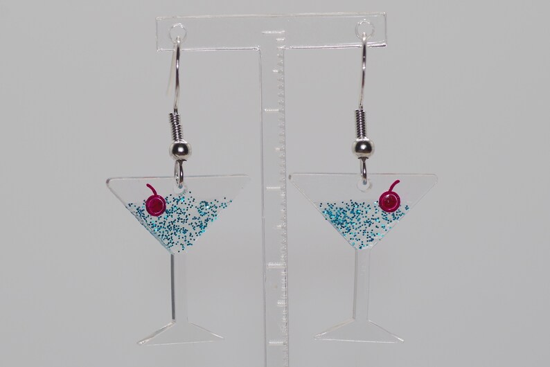 May include: A pair of silver dangle earrings with a clear glass martini glass charm filled with blue glitter and a red cherry on top.