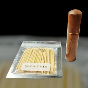 May include: A clear plastic bag filled with wooden toothpicks. The toothpicks are labeled "Bourbon Pickin Sticks". A wooden container with a gold band is next to the bag.