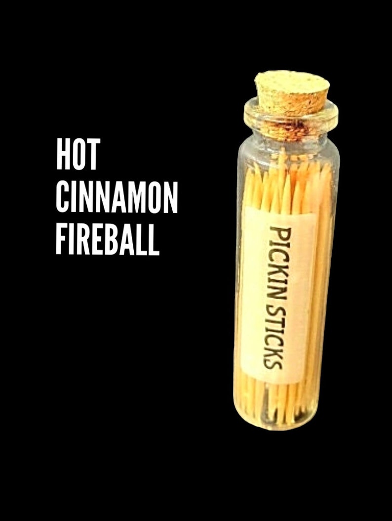 Może przedstawiać: Mały szklany słoik wypełniony drewnianymi wykałaczkami. Słoik ma korkową pokrywkę i etykietę z napisem "Pickin Sticks". Tekst "HOT CINNAMON FIREBALL" jest napisany białymi literami na czarnym tle.