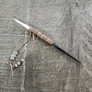 May include: A handmade spear with a white tip, a brown wrapped handle, and a black shaft. A leather cord with silver beads is attached to the handle. The spear is lying on a gray, textured surface.