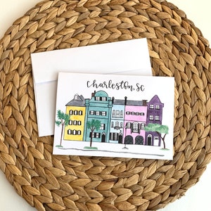 Pode incluir: Um cartão de felicitações com uma ilustração de aquarela de casas coloridas em Charleston, Carolina do Sul. O texto "Charleston, SC" está escrito em itálico no topo do cartão.