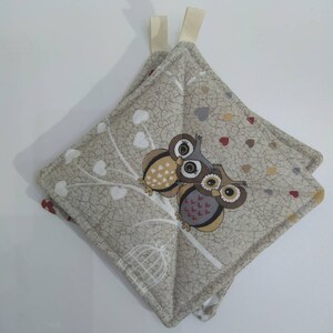 Może przedstawiać: Dwa beżowe uchwyty do garnków z wzorem białych serc i brązowych sów. Sowy siedzą na gałęzi z klatką dla ptaków w tle.